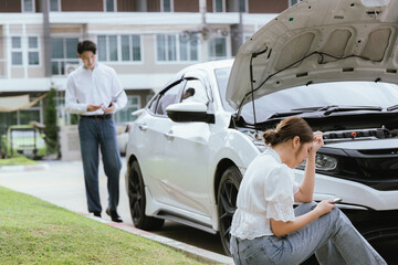 A young Caucasian man and an Asian woman inspect a damaged car after an auto accident. The...
