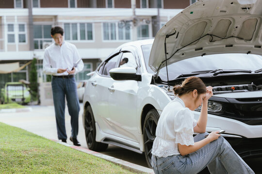 A young Caucasian man and an Asian woman inspect a damaged car after an auto accident. The insurance agent examines the vehicle, writing a report while gathering evidence for the claim investigation a