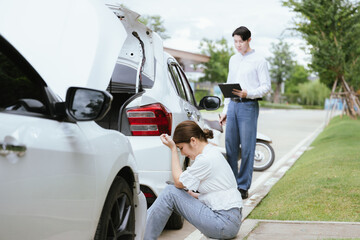 A young Caucasian man and an Asian woman inspect a damaged car after an auto accident. The insurance agent examines the vehicle, writing a report while gathering evidence for the claim investigation a