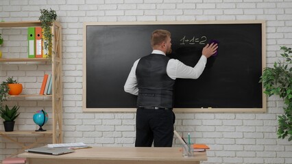 Man college teacher in classroom in front of chalkboard explaining lesson for students, writing simple math tasks. Education concept.