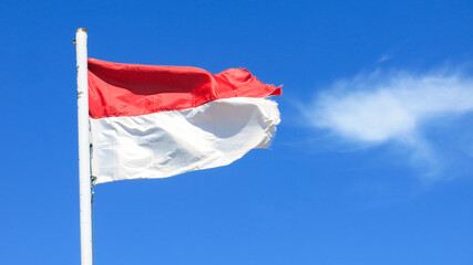 The red and white Indonesia national flag is waving againts clear bright blue sky. little soft white cloud behind.