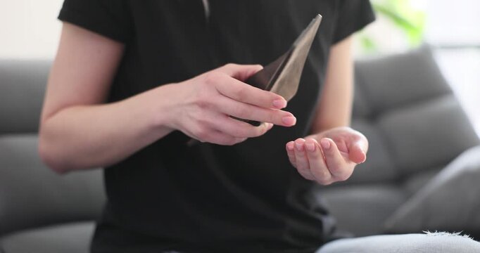 Woman shakes out last coins from empty wallet sitting on sofa. Lady short in money seeks cash in purse at home. Problem of poverty