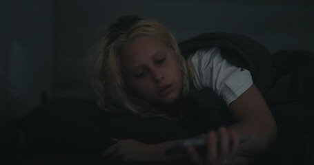 A blonde girl in a white T-shirt wakes up abruptly and looks anxiously at the phone at night