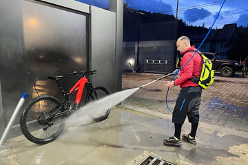 Cyclist using high-pressure water spray to clean electric mountain bike on outdoor washing station.