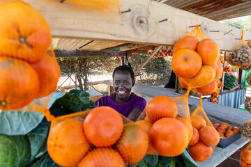 Happy African street vendor hawker village , woman with short hair , shack with vegetables and...