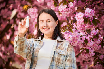 Woman allergic using medical nasal drops, suffering from seasonal allergy in blossoming garden. Smiling woman holding nasal spray near blooming tree. Spring allergy concept.