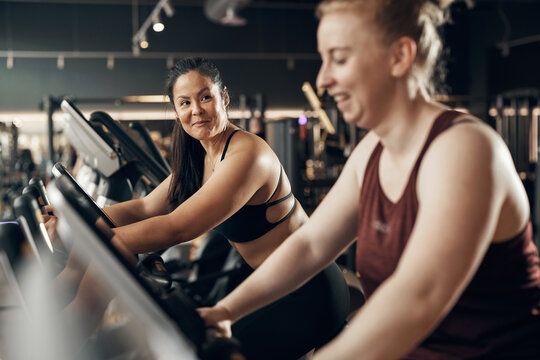 Diverse smiling young woman and a female friend in sportswear riding on stationary bikes during a cardio workout session together at the gym - Powered by Adobe