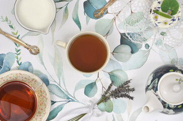 Two cups of tea, Tea pot, milk, cupcake and two spoons on a table painted with green leaves. Top view, British style.