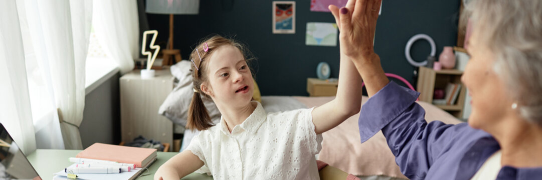 Header image of young girl with Down syndrome giving high five to grandmother while doing homework together and enjoying studying - Powered by Adobe