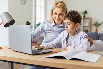 Positive mother assisting attentive schoolboy with homework using a laptop for remote learning at home. Family cooperation ensures effective education and support during study time.