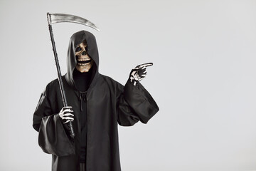 Male person in Halloween grim reaper death costume holding scythe in black cloak with hood pointing...
