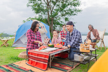Asian family enjoying lakeside camping picnic. Parents and child prepare watermelon, grandparents relax nearby. Blue tent, colorful blanket, and camping gear create cozy outdoor scene.