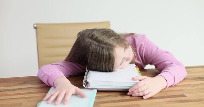 Junior schoolgirl clearly exhausted rests head on notebook. Small child with loose hair struggles to complete homework with tired eyes barely open