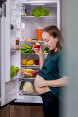 Pregnant woman is eating an apple near the refrigerator