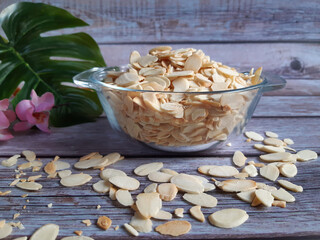 Clear bowl of sliced almonds on a wooden surface, with scattered nuts, a green leaf, and a pink flower behind, perfect for showcasing healthy food ideas.