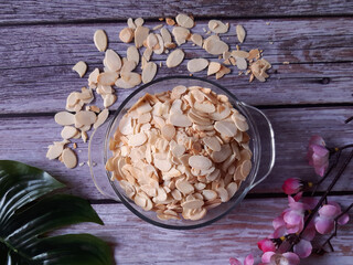 Bowl filled with almond slices resting on wood, with additional almonds scattered around and a green leaf, plus a pink flower, highlighting healthy food concepts.