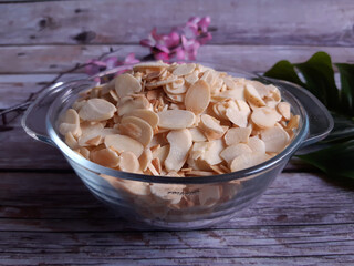 Glass bowl of almond slices on a wooden surface, surrounded by scattered almonds, with a green leaf and pink flower in the background, ideal for natural food displays.