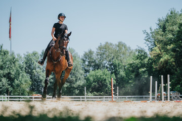 A rider wearing a helmet practicing equestrian skills on a horse in an outdoor arena with jumps on...