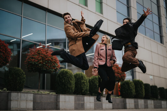 Three young professionals in smart casual wear are leaping joyfully in the air outside a contemporary office setting, displaying a sense of freedom and happiness.