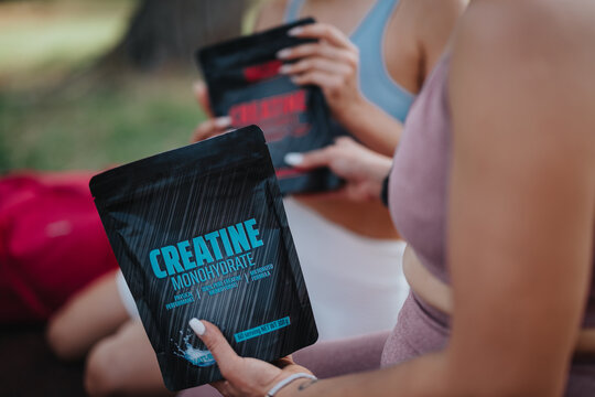 Close-up of women holding creatine monohydrate supplement packs during outdoor fitness session in park.