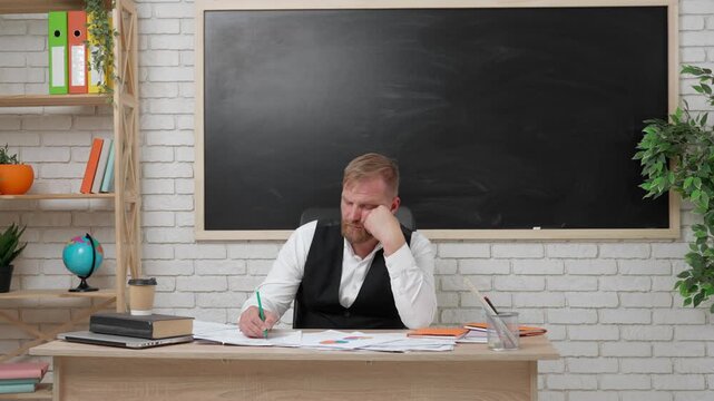 Man college teacher sit at desk in classroom in front of chalkboard, checks exam test papers, tired expression, falling asleep. Education concept.