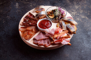 Snacks on wooden utensils. Sliced bacon and hunting sausages. Small snacks for beer in the form of fish on bread. Food on a dark texture background.