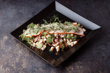 Summer salad with pieces of bacon, fresh cucumbers, cheese cubes and mushrooms. Green salad on a black plate on a dark background. Free space for an inscription. View from above.