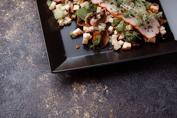 Summer salad with pieces of bacon, fresh cucumbers, cheese cubes and mushrooms. Green salad on a black plate on a dark background. Free space for an inscription. View from above.