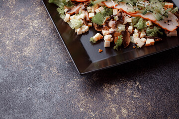 Summer salad with pieces of bacon, fresh cucumbers, cheese cubes and mushrooms. Green salad on a black plate on a dark background. Free space for an inscription. View from above.