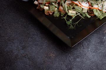 Summer salad with pieces of bacon, fresh cucumbers, cheese cubes and mushrooms. Green salad on a black plate on a dark background. Free space for an inscription. View from above.