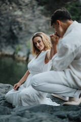 A romantic couple dressed in white clothes, sharing a tender moment near a rocky terrain by the water, conveying love and connection.