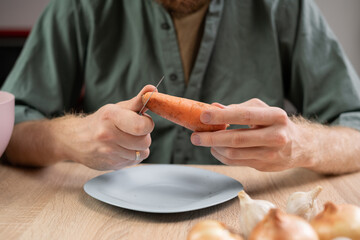man peeling a carrot with knife, cooking at the wooden table in the kitchen