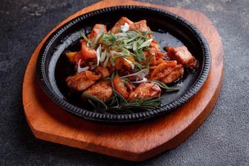 Hot frying pan with pieces of fried meat in caramelized sauce. Cooked and fresh vegetables as garnish and garnish.