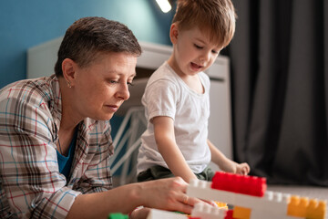 Obraz premium Middle-aged mother helps son build block house on floor