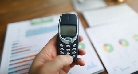 In the hands of an old model of a mobile phone against the background of reports on the table, close-up, blurry. Corporate cell phone