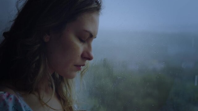 Natural beauty woman crying standing looking at window rain drops falling dark rainy weather mood depression sadness loneliness emotion. Adult Girl sad tear-stained face eyes closed. video footage 4k