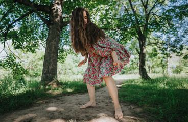 A girl in a floral dress performing modern dance outdoors on a dirt path, surrounded by lush green trees and sunlight.