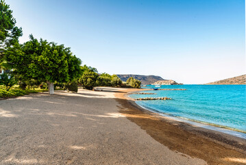 Naklejka premium Coastal scene featuring empty beach with mix of sand and pebbles along shoreline. Calm, blue waters of sea gently meet shore, while lush green line of trees and vegetation creates natural border 