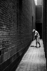 A man sweeps a narrow alleyway bordered by textured brick walls.