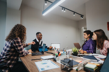 A dynamic scene depicts a multicultural business team actively discussing strategies and project details around a cluttered office table, enhancing team collaboration.