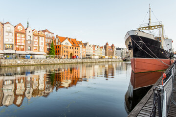  A historic black and red ship is docked along the Motlawa River in Gdansk, Poland, with picturesque buildings reflected in the water.