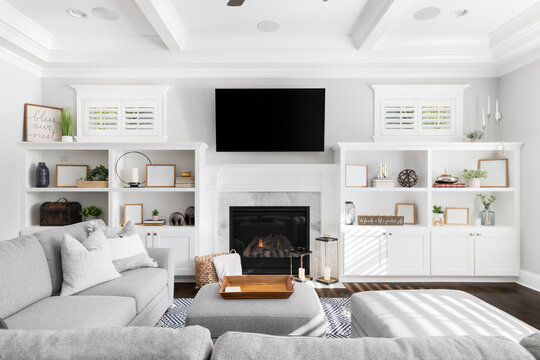 A living room detail with a large grey couch looking towards built-in shelving, a television over a fireplace, and a coffered ceiling above. Picture frames are blank.