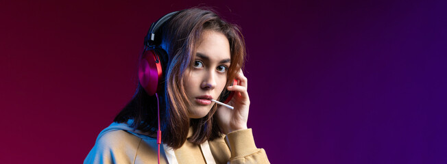 Young beautiful fashionable hipster girl dressed in a hoodie  listening to music in red headphones in a studio red dramatic background