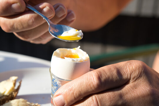A person is scooping a yellow egg yolk from soft boiled egg with shell during breakfast time.