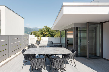 External balcony of villa modena with a small living room composed of a sofa, two armchairs and a table with chairs. In the background Swiss hills
