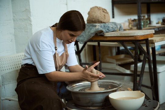 Young woman in apron making ceramic pot on potters wheel