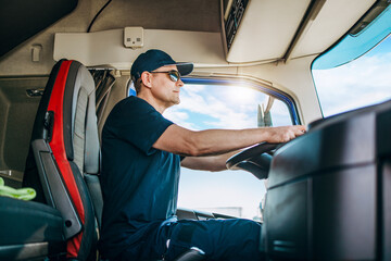 Handsome experienced male truck driver with a hat sitting and driving his truck. Professional transportation and truck drivers concept. Side view.