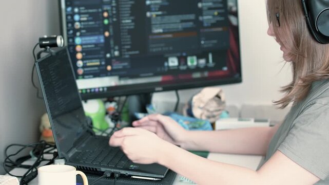 Working at home on a laptop. A young caucasian girl working on a notebook with second display. Typing text on a keyboard while having friendly chat with someone. Online meeting. 