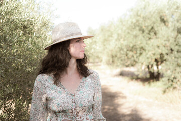 Chica joven y guapa se pasea y se divierte tranquila entre los olivos de un olivar en verano en una tarde soleada con vestido y sombrero