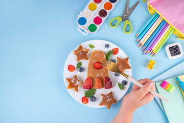 Obraz premium Back to school, school kids breakfast with creative shaped pancakes and fruit berries, on light blue background with school supplies, top view copy space. Kids hands eating pancakes